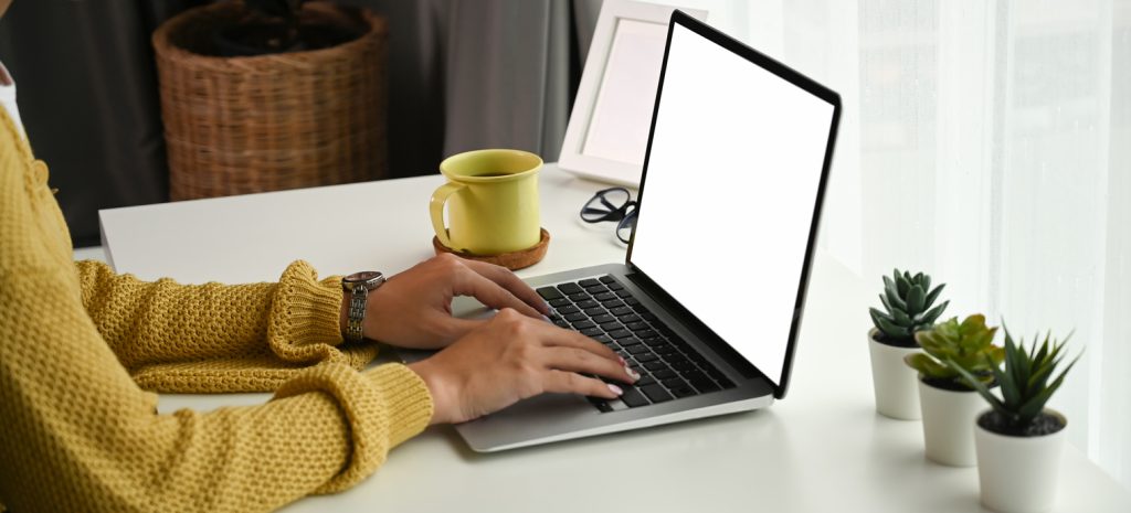 Cropped shot of of young woman typing on laptop with blank white desktop screen while sitting at her workspace.