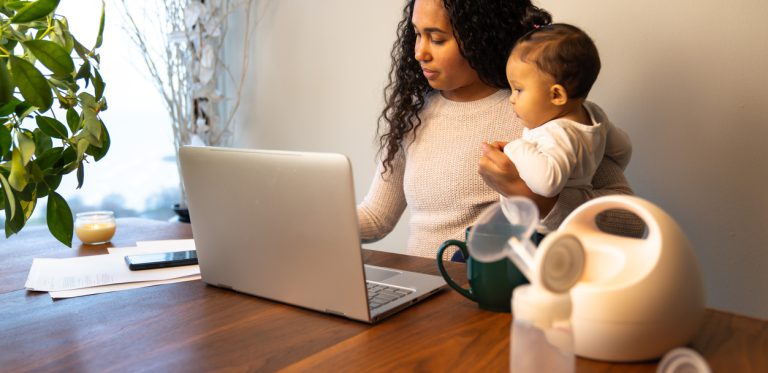 mother holds her daughter while taking notes at her dining table serving as a temporary remote work from home station with breast pump in foreground.